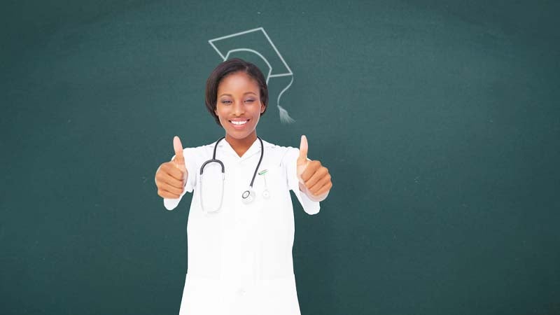 Graduate who achieved a degree in nursing holding two thumbs up and standing against a chalkboard